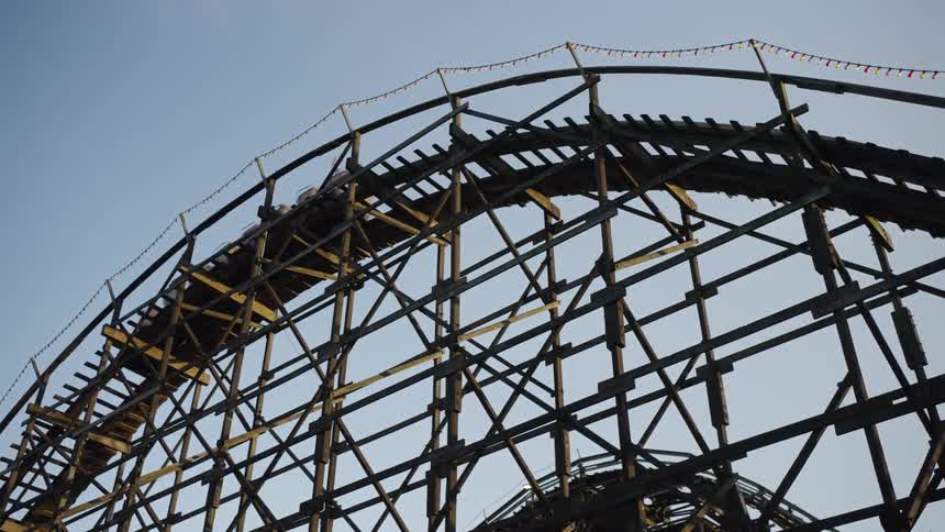 A wooden roller coaster rises steeply beside leafy trees under a clear blue sky