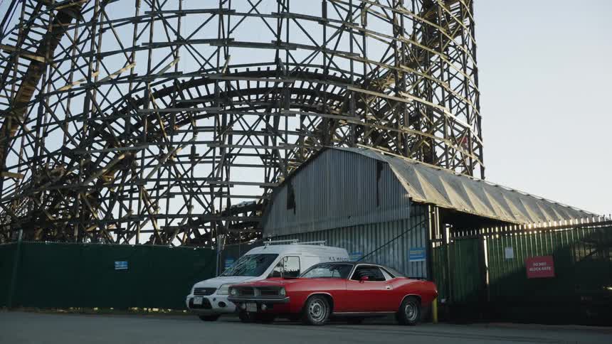Roller coaster structure rises behind a warehouse with a vintage car parked in front