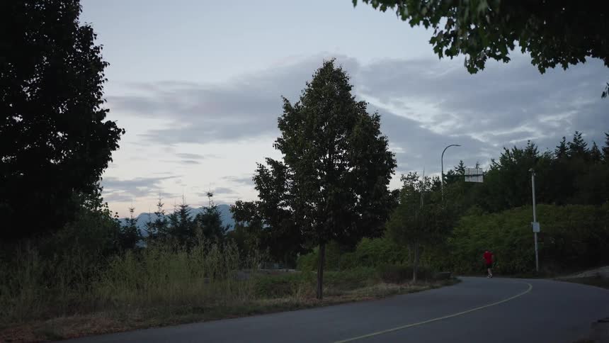 A man wears a red tee shirt while jogging on a forested road at dusk