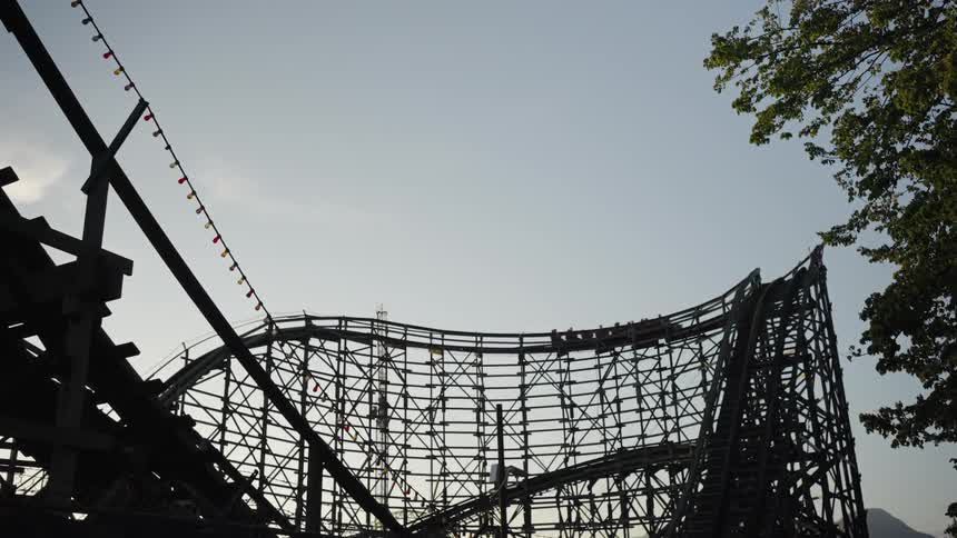 The video shows a lowangle shot of a classic wooden roller coaster the camera follows a coaster car as it slowly ascends a chain hill silhouetted against a bright sky