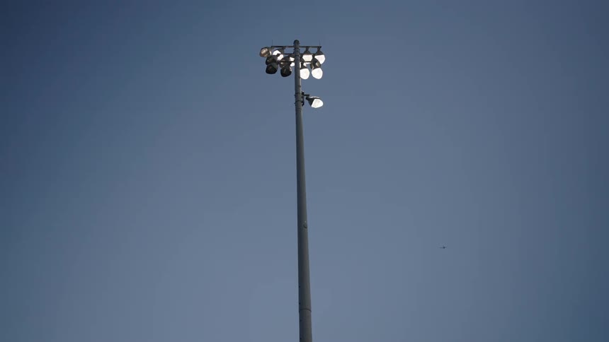 A tall stadium floodlight stands against a clear twilight sky with a distant airplane barely visible