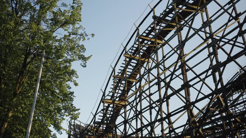 A wooden roller coaster with riders rises steeply beside leafy trees under a clear blue sky