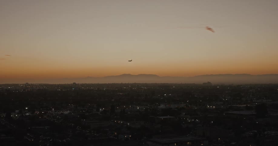 A plane glides across a glowing orange sky at dusk above a sprawling city with twinkling lights