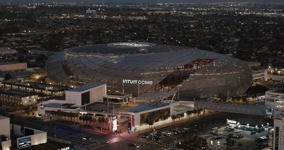 A sports stadium with a round roof in the city at night with a busy street beside it as cars go by