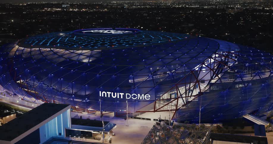The video shows an exterior shot of a large modern stadium at night with a prominent intuit dome sign the building is illuminated with blue lights and the camera pulls back to reveal the surrounding cityscape