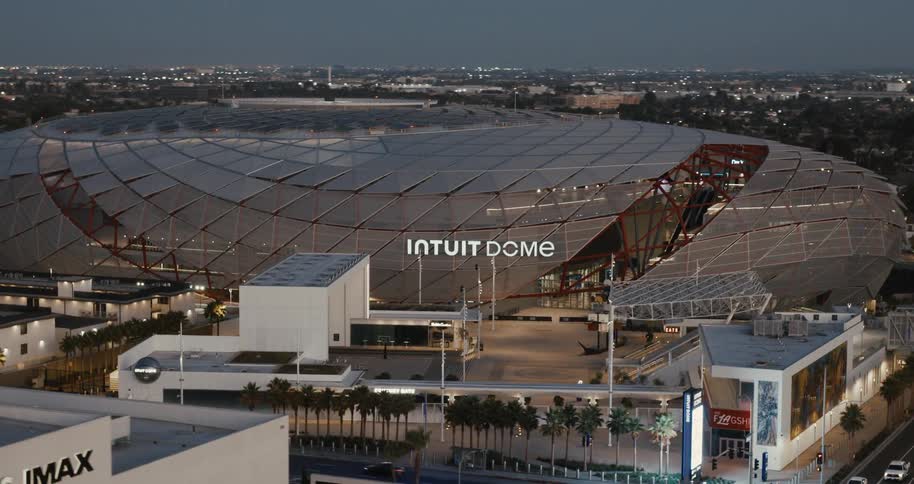 Intuit dome stadium appears lit up at dusk in the middle of the city