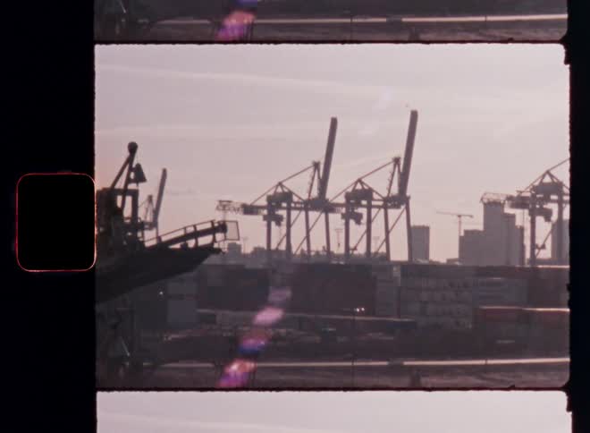A large cargo ship is docked in a port with tall cranes behind it all in silhouette against a hazy orange and pink sunset