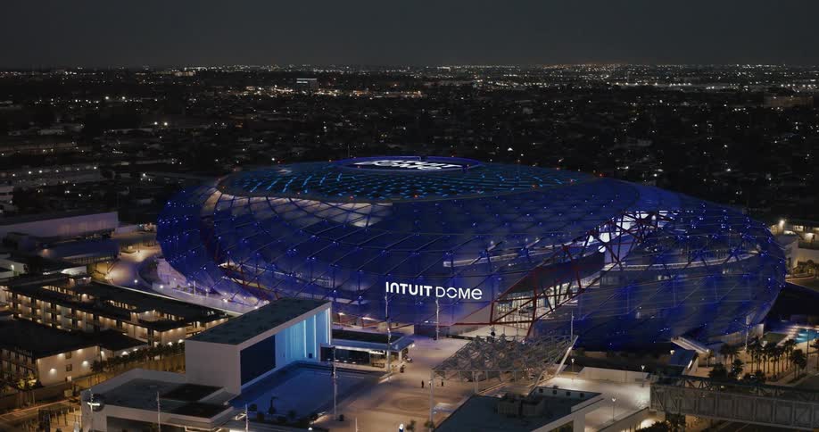 An aerial view of a large modern stadium with a curved blue dome is brightly lit in the middle of a dark city at night