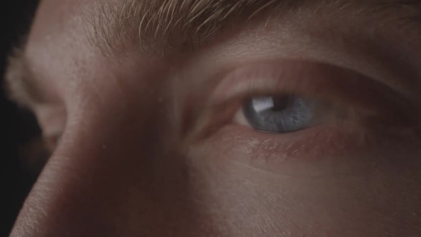 A closeup reveals the intense blue eye of a person gazing forward in soft lighting