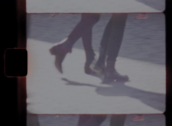 A vintage film frame captures people walking focusing on their legs and casting long shadows
