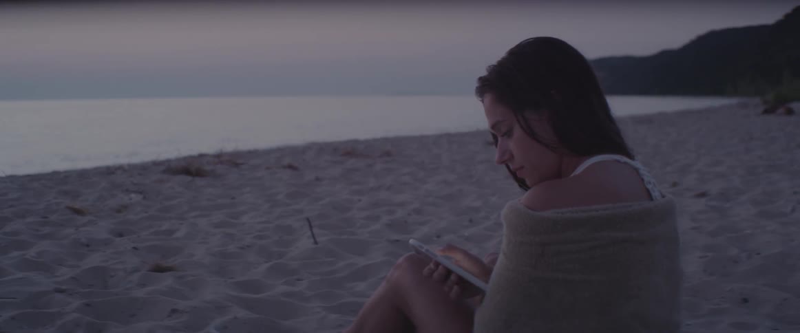 A woman uses her phone while sitting on a beach at sunset