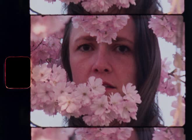 Woman stands between the branches of a tree and stares past its pink flowers surrounding her body and head with long brown hair on retro 8mm film footage with arty avantgarde vibes