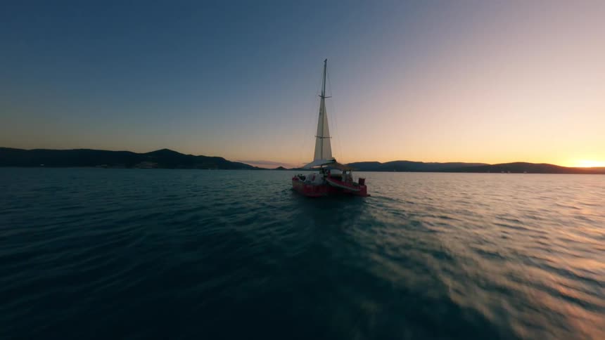 A sailboat sailing in the ocean with the shore and the orange setting sun in the background