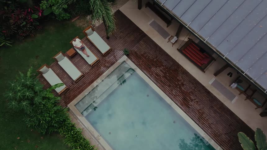 Aerial of a man using a laptop computer on a chair beside a pool outdoors in a yard during daytime