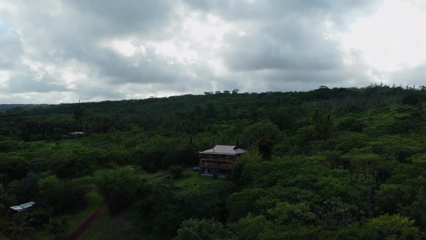 A small house sits nestled in dense tropical greenery beneath a sky filled with dramatic gray clouds