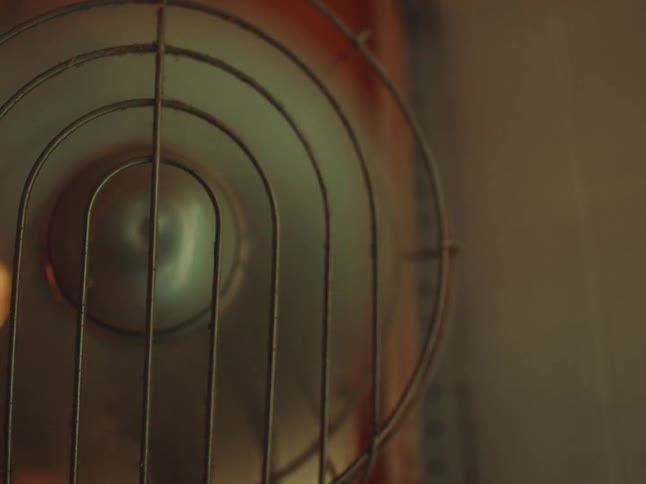 A closeup reveals the metallic spiral guard of a vintage fan softly lit in warm tones
