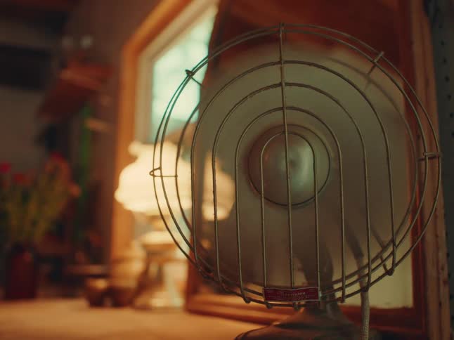 A standing fan blowing at full speed inside a room positioned in front of a mirror with a lampshade on the side casting light