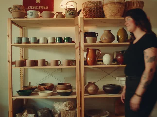 A female walks past wooden shelves display in a curated collection of ceramic mugs bowls and pottery in a warm homey studio