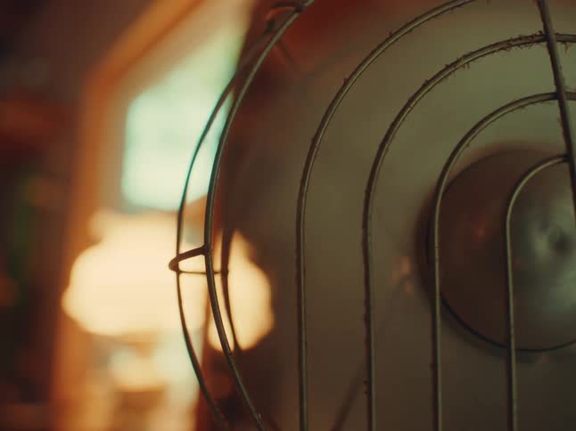 A close view of an old metal fan with a safety cage is in focus with a blurry golden light visible in the background