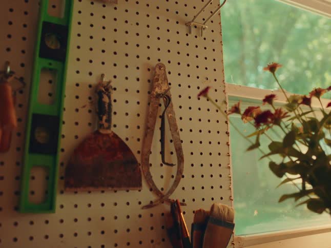 The camera slowly zooms in on a white pegboard wall with a green level a rusty trowel and other tools hanging on it with a vase of red flowers on a table in front of a window