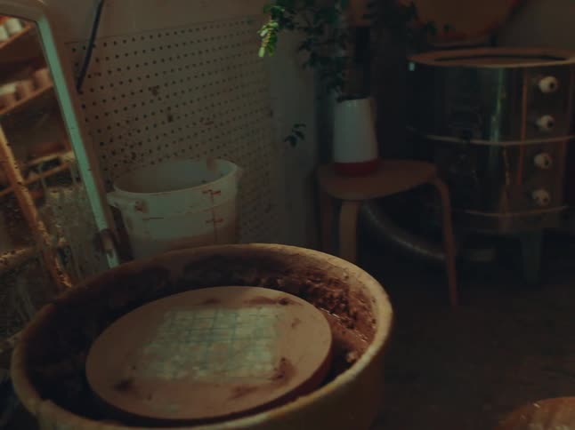A slow panning shot across a rustic cluttered pottery studio revealing a wooden shelf filled with various ceramic pieces and raw clay a potters wheel and other tools of the trade
