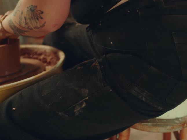 A tattooed individual works with clay on a pottery wheel their clothes streaked with mud in a cozy studio