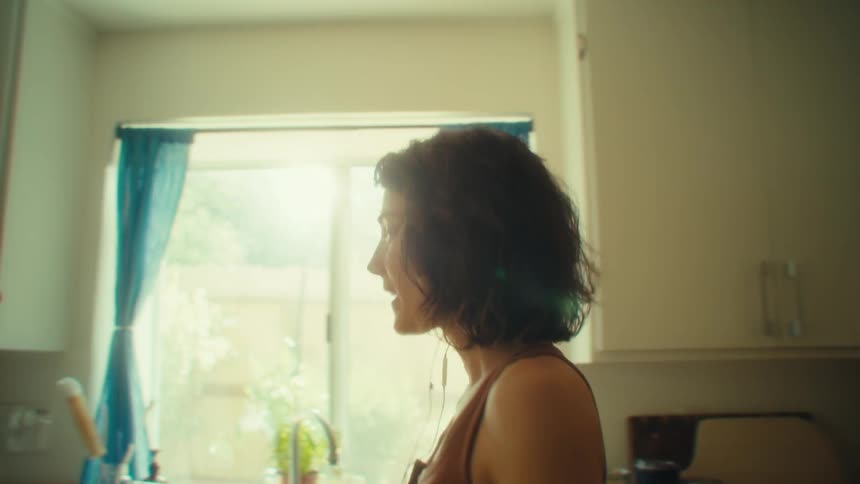 A young woman in the kitchen at midday wearing wired earbuds singing and dancing while doing her chores