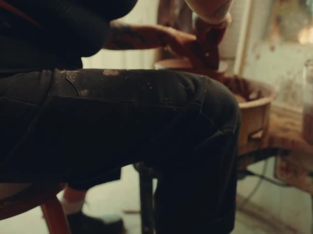A woman with a tattoo on her arm sits facing a mirror inside a studio at midday using a pottery wheel to make a clay pot