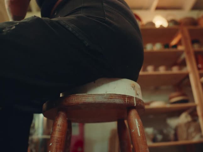 A closeup of a wooden stool beneath the seated person with pottery shelves blurred in the background