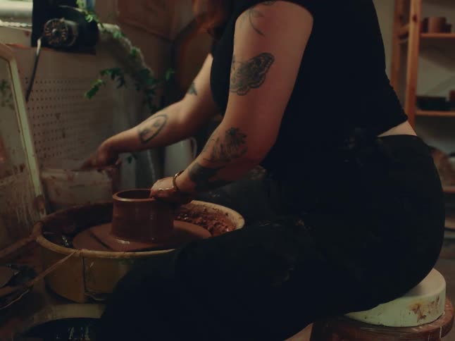 A tattooed individual shapes wet clay on a pottery wheel in a warmly lit studio filled with tools and plants