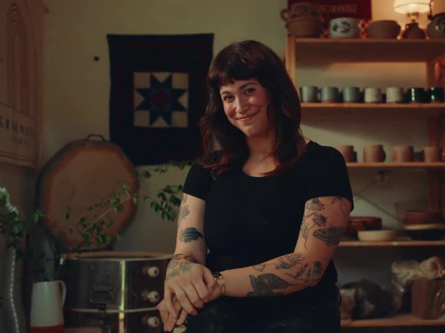 A woman with tattoos sits confidently in a cozy pottery studio surrounded by handmade ceramics and warm lighting