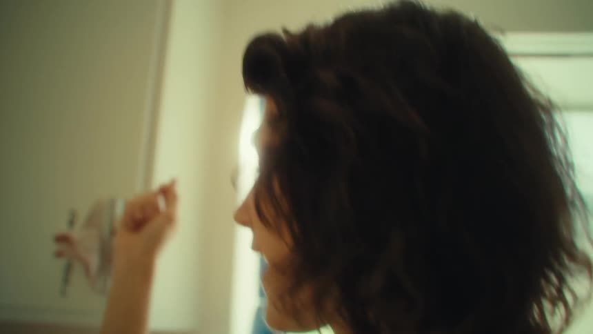 A woman in a rustcolored tank top moves through a bright kitchen holding a glass bathed in soft natural light