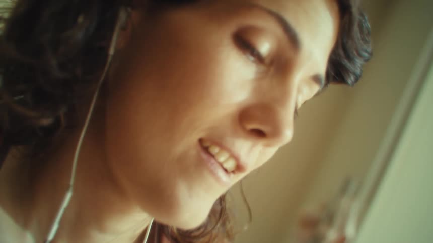 A young woman wearing earbuds in the kitchen laughing and having fun while doing chores at midday