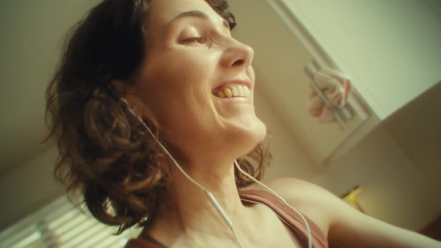 A woman with brown wavy hair and earbuds in her ears sings while washing a large bowl in a sunlit kitchen