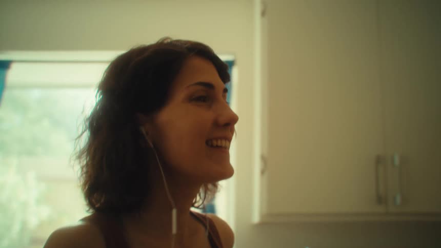 A woman with short dark hair wearing a brown tank top and wired headphones smiles while she dances through a sunlit kitchen with a clear glass in her hand