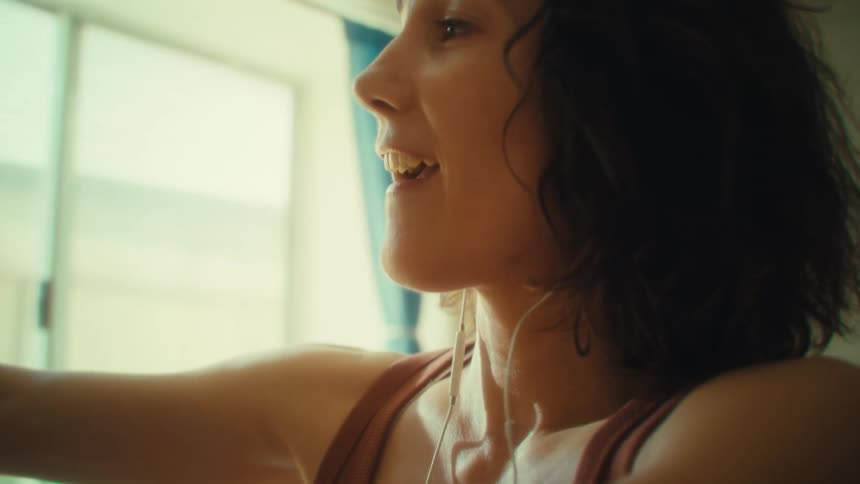 A woman with earbuds smiles and sings while reaching up to put a glass away in a sunlit kitchen with bright blue curtains