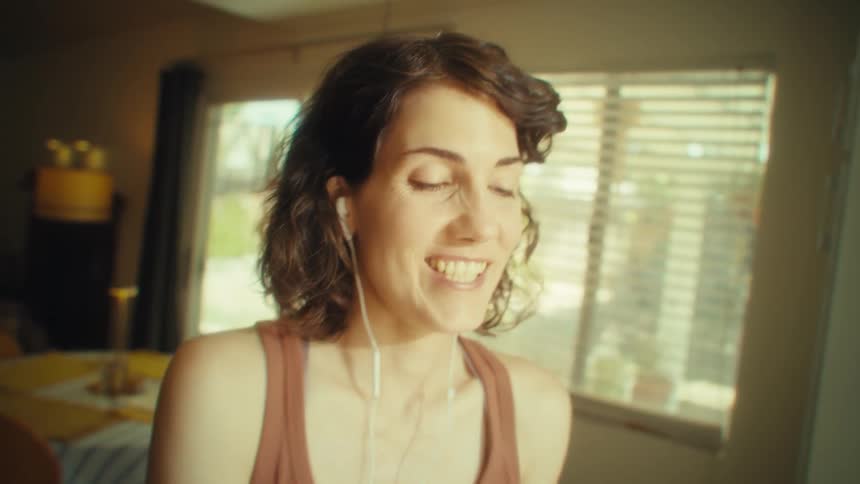 A woman in a sunlit room smiles and sings with closed eyes while listening to music through white earphones