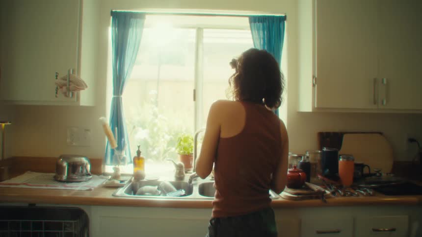 Woman in a brown tank top washes dishes in a kitchen lit by a window with blue curtains