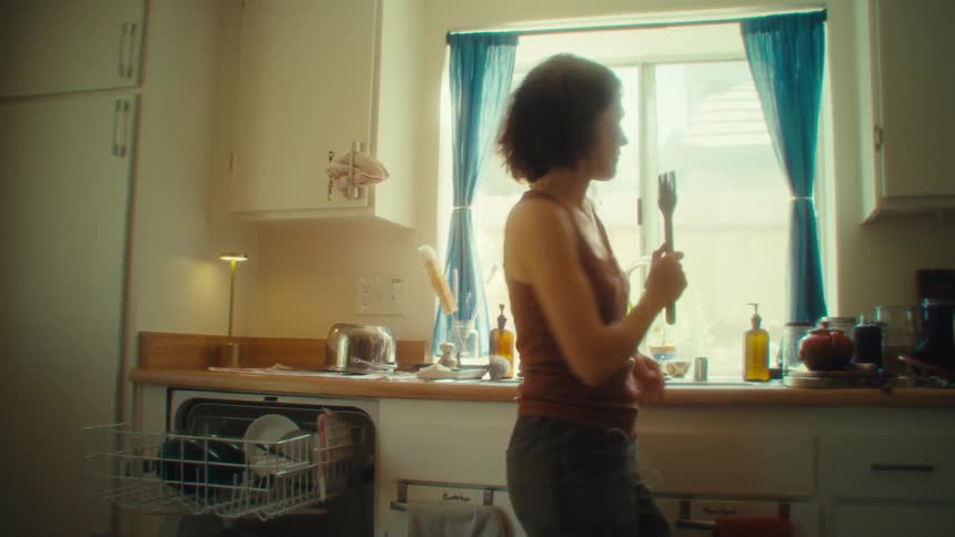 A woman with a silhouette against a bright sunlit window dances and moves casually in a home kitchen
