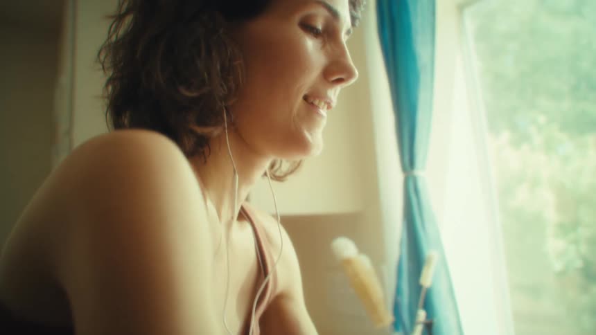 A woman smiles softly as she listens to something through earbuds and washes dishes in a warmly lit kitchen