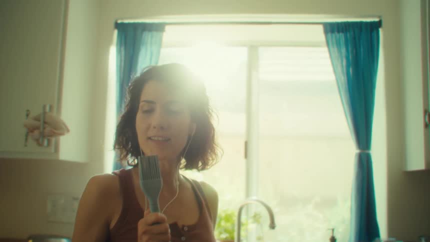 A young woman spins and dances as she sings in the kitchen while doing dishes during daytime