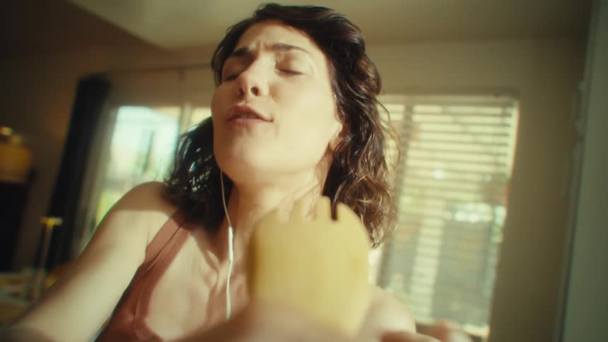 A young woman in her kitchen at midday wearing earbuds and singing into a wooden spoon as if it were a microphone having fun