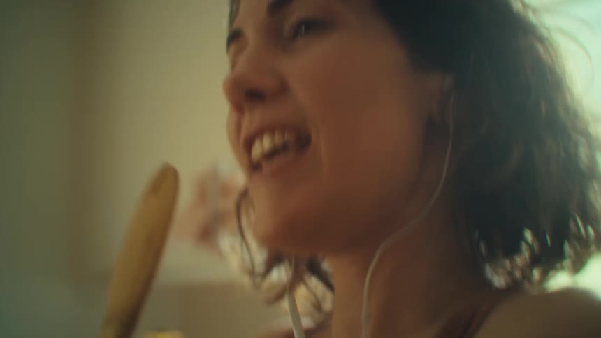 Close up portrait of a young woman singing with a utensil while listening to music with earphones in the kitchen during daytime
