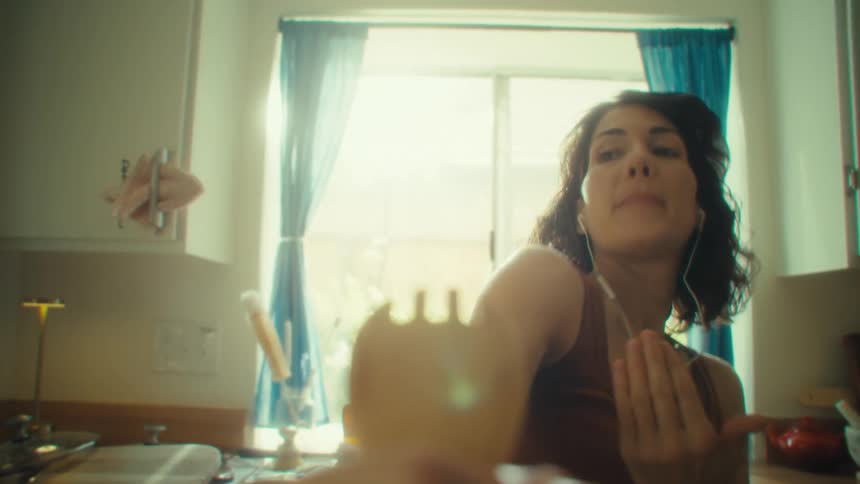 A woman sings passionately into a wooden utensil while wearing earphones in a sunlit kitchen
