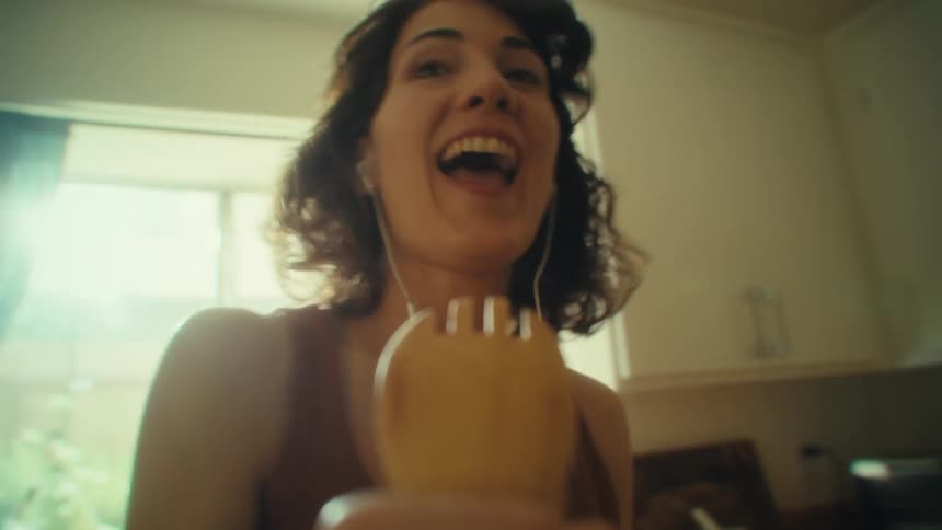 A woman sings and dances while energetically pointing a finger in a sunlit kitchen while she holds a large wooden fork