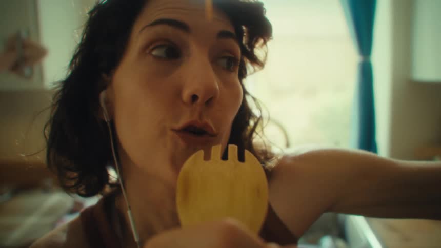 A young woman in the kitchen at midday wearing wired earbuds and singing into a spoon having fun