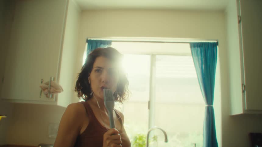 A female sings into a kitchen brush while dancing in a kitchen on a sunny day