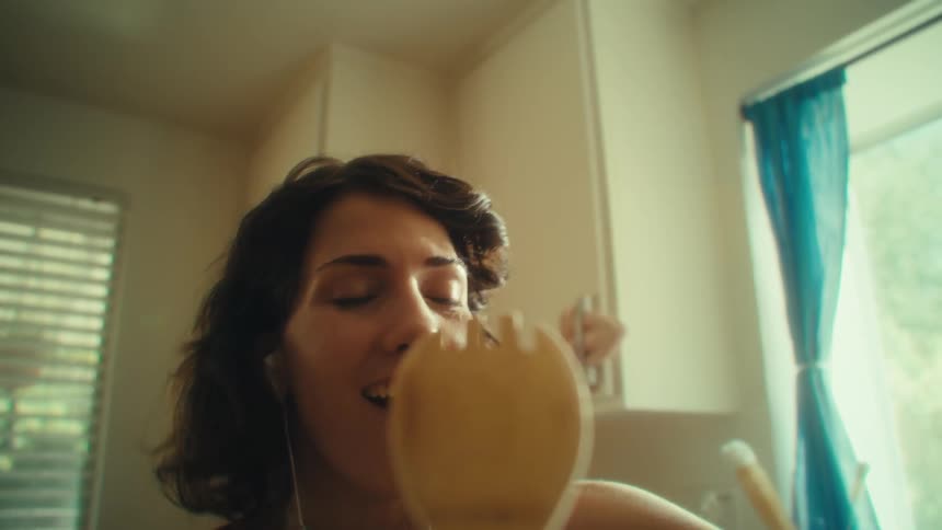 A young woman in her kitchen at midday wearing earbuds and singing into a wooden spoon as if it were a microphone