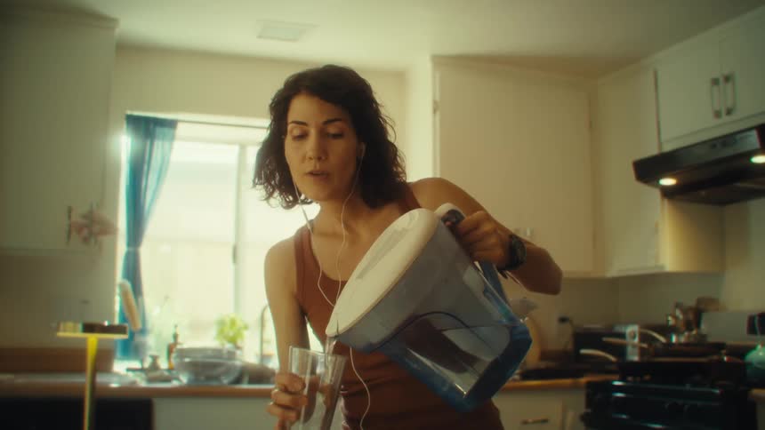 A woman pours water into a glass while listening to music in her kitchen