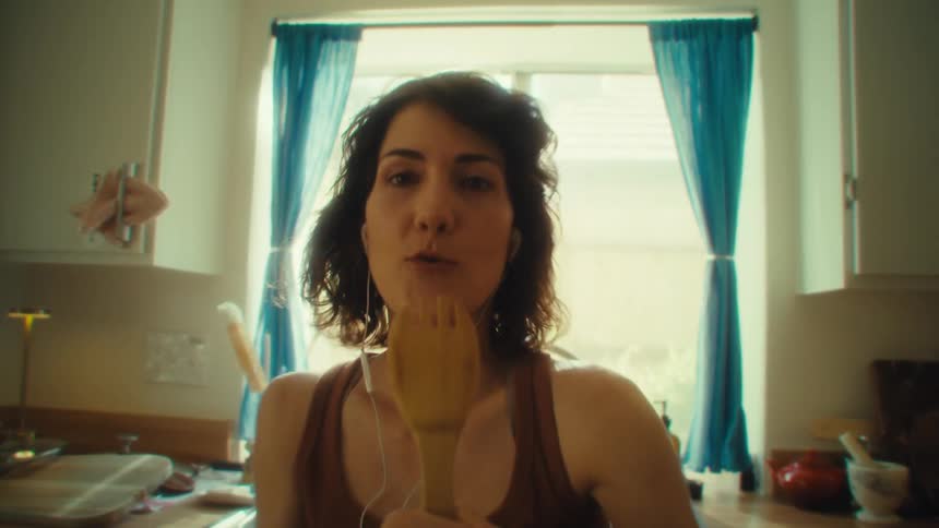 A woman listens to music and sings into a wooden spoon in a sunlit kitchen with blue curtains